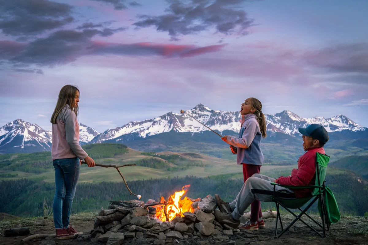 Children and a father figure gathered around a campfire, roasting marshmallows with snowy mountains in the background. The scene radiates warmth, togetherness, and a spirit of adventure, reflecting cherished family moments in nature.