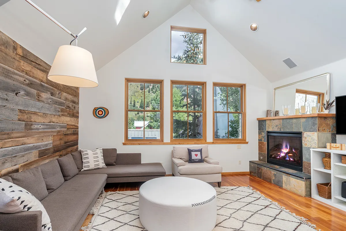 A welcoming living room with a cozy sofa positioned around a gas fireplace, large windows flooding the space with natural light, and a round coffee table at the center. The soaring ceiling and expansive windows capture the mountain scenery beyond, enhancing the sense of openness and connection to nature.