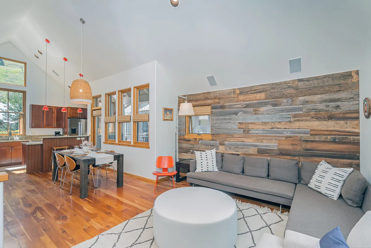 A stylish, inviting space featuring a wooden accent wall, modern couch, and a set dining table under charming pendant lights. The room's layout emphasizes communal gathering, with sightlines to both living and dining areas for seamless interaction.