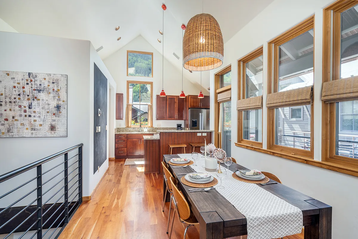 The image showcases a beautifully lit dining area and modern kitchen, featuring elegant cabinetry, a chic light fixture, and expansive windows that fill the space with natural light. The scene is set for warm family meals or lively gatherings with friends, creating an inviting and comfortable atmosphere.