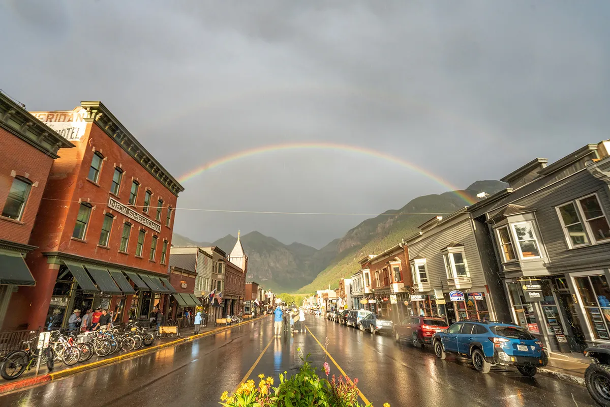 A lively Main Street scene featuring historic brick buildings, wet street reflections after a light rain, and a cheerful rainbow arching overhead. Local boutiques and outdoor cafes are visible, inviting exploration and relaxation.
