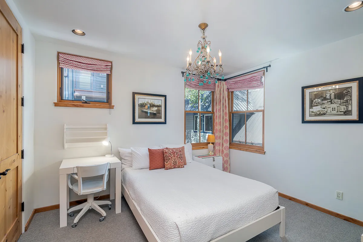A stylish bedroom featuring a comfortable bed with soft bedding, an elegant chandelier, a dedicated desk space with a chair, tasteful artwork on the walls, and gentle, soothing lighting. The space feels both relaxing and functional.