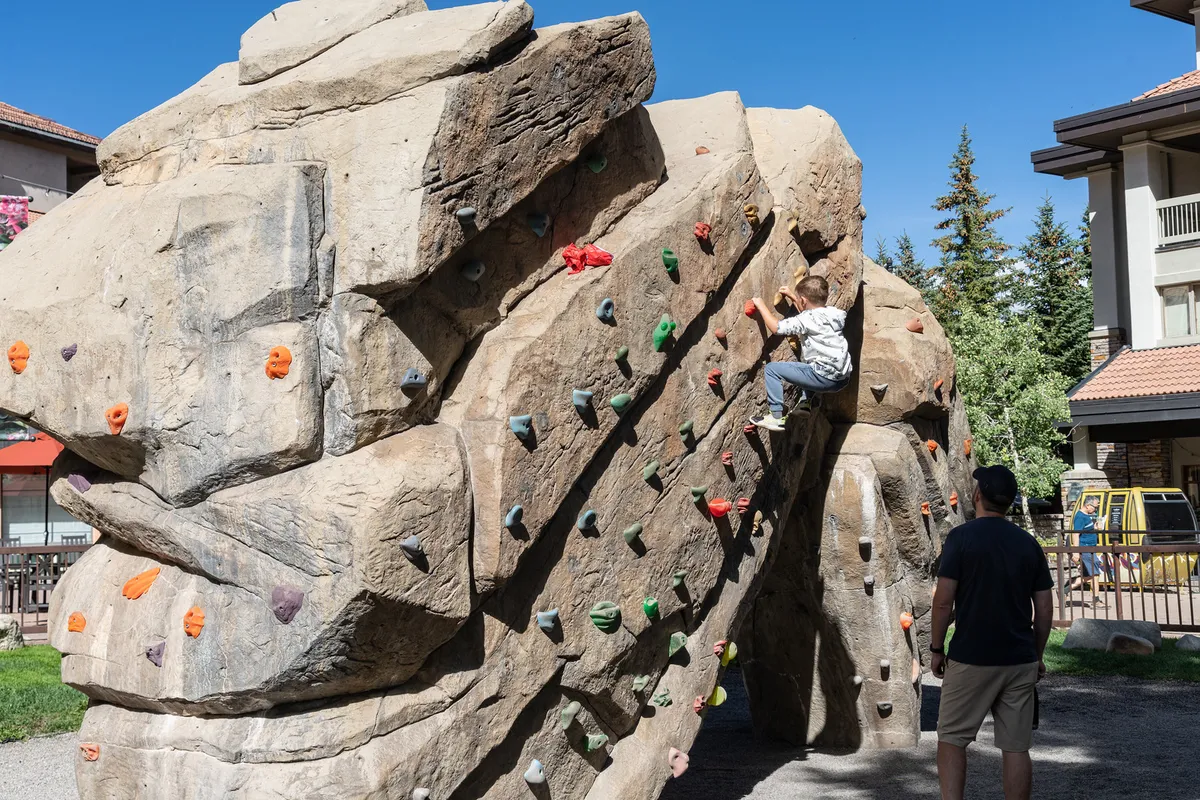 A child climbing on a rock structure under the watchful eye of an adult, set in an outdoor recreational area. The backdrop suggests a safe, playful environment ideal for children to explore and develop new skills while being supervised.