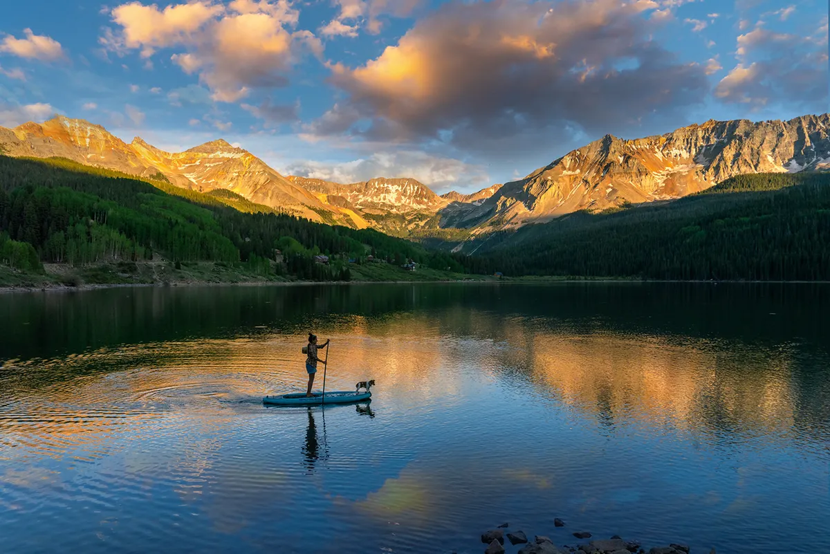 A paddleboarder and their dog glide peacefully over a tranquil river or lake, with the still water reflecting the distant mountain range. The golden light of sunset enhances the serene, adventurous ambiance.