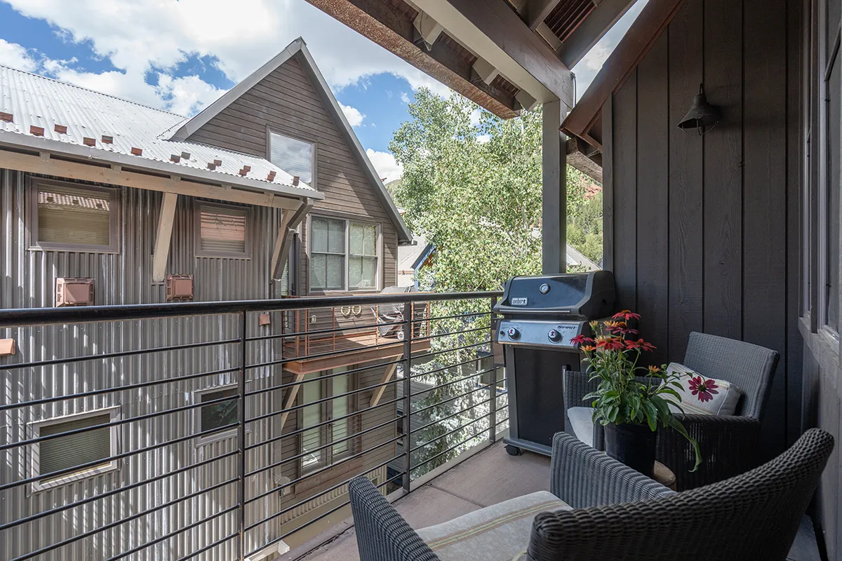 This image captures a private balcony scene featuring a modern railing, an inviting grill, a seating area, and a planter brimming with colorful flowers. The balcony is bathed in natural light and offers a tranquil view, hinting at mountain scenery beyond. The space feels ready for both quiet mornings and lively gatherings with friends.
