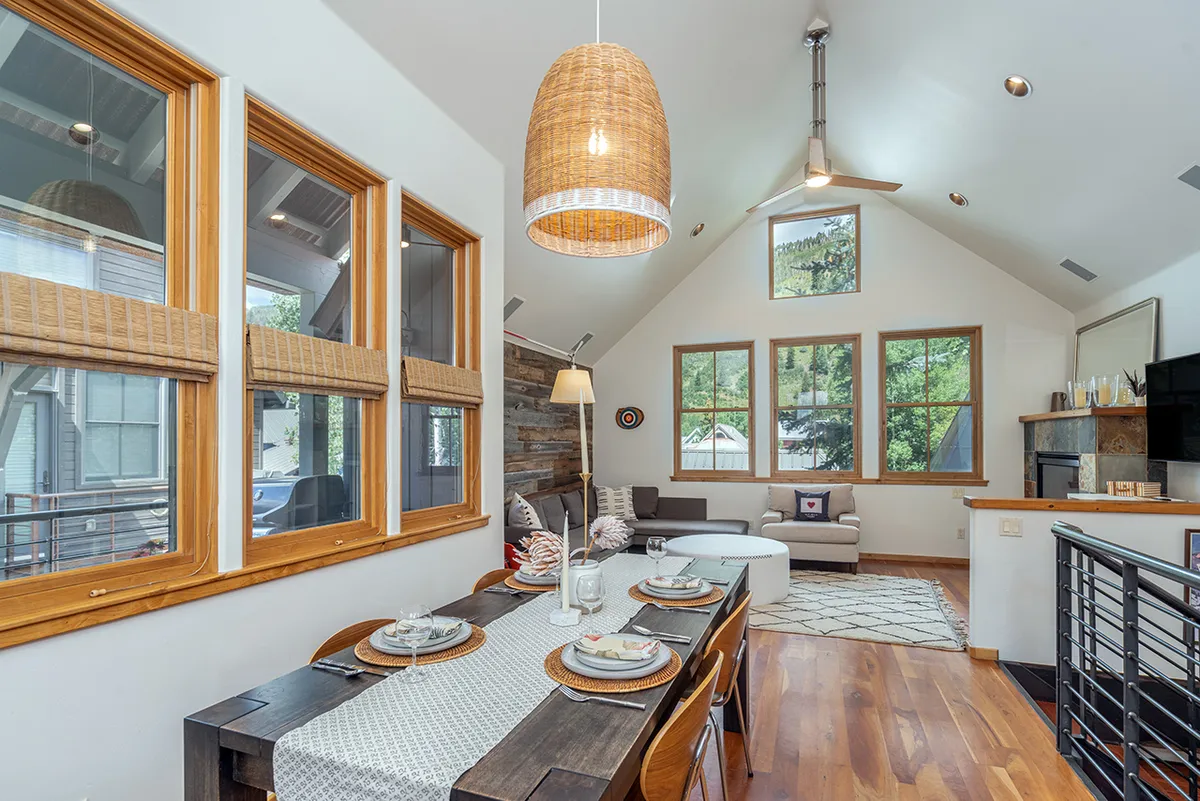 A modern, cozy dining area with a large table, pendant light, and big windows that fill the space with natural light. The living area in the background suggests space for gatherings and relaxation after a day of adventure.