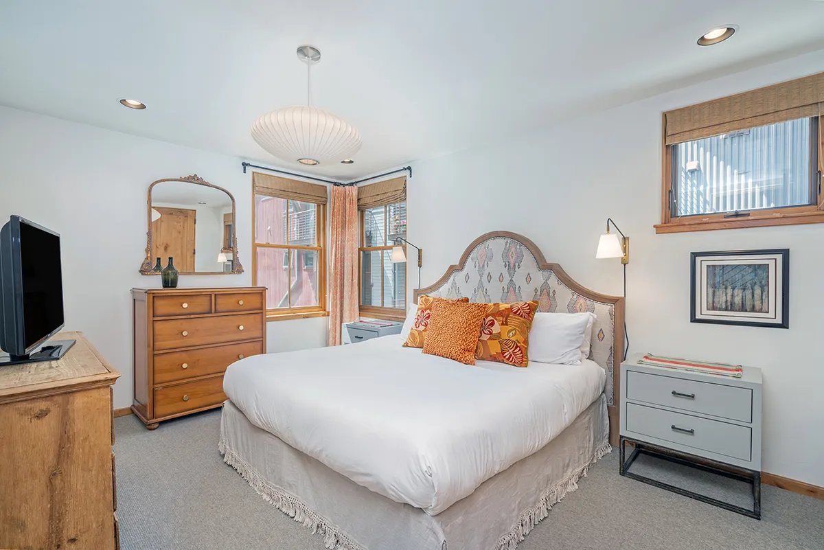 A beautifully arranged bedroom with a plush bed adorned with decorative pillows, a chest of drawers, a large mirror, a television, and a window letting in natural light. The ambiance is cozy, welcoming, and restful, ideal for relaxation and privacy.