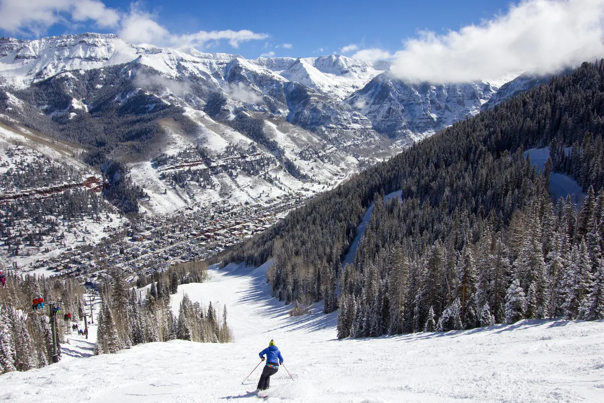 A skier is descending a powdery slope with snow-covered mountains and dense evergreens in the background. Below the slopes, a small town nestles into the valley, illustrating the close connection between mountain adventure and community life.