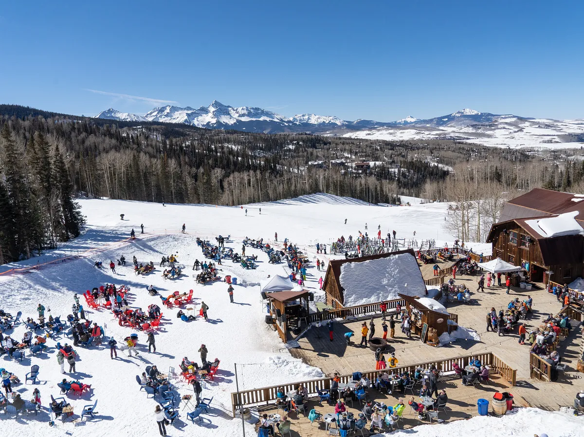 A bustling mountain scene with people socializing on a wooden deck, skiers and snowboarders on the snowy slopes, and a picturesque mountain backdrop under bright sunshine. The festive energy is palpable as people gather for Après-ski moments.
