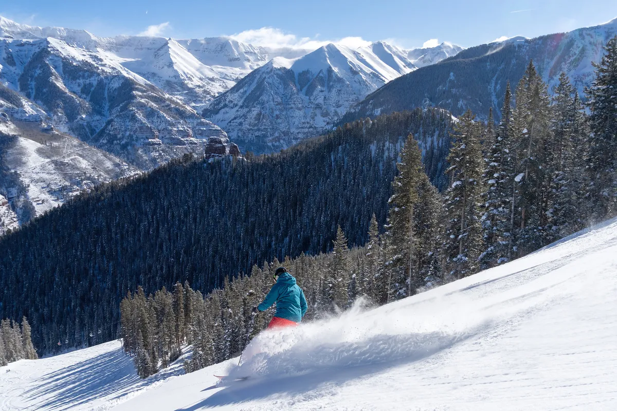 A skier carves through pristine snow on a mountain slope, surrounded by pine trees and snow-capped peaks under a clear sky. The foreground captures the thrill of skiing, while the background showcases the breathtaking mountain scenery Telluride is known for.
