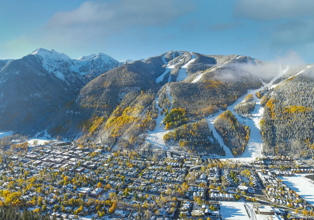 A panoramic view of Telluride with snow-capped mountains framing the town area below and colorful autumn foliage in the foreground. The image captures both natural beauty and the proximity of town amenities, with hints of ski slopes and festival venues.