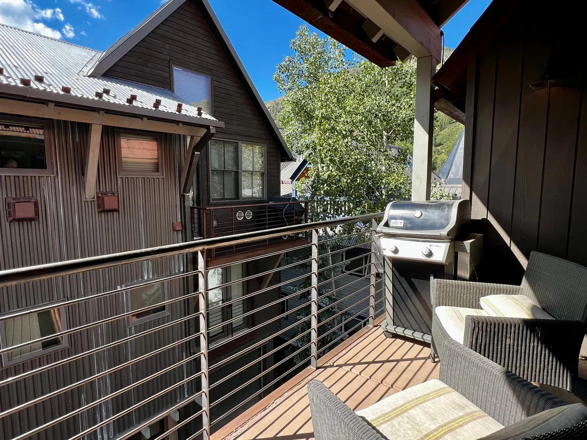 A relaxed outdoor setting on a balcony with a prominent grill, comfortable seating, and a lush view of surrounding treetops. The scene suggests a casual gathering spot for barbecues or tranquil mornings with coffee, set against a backdrop of nature.